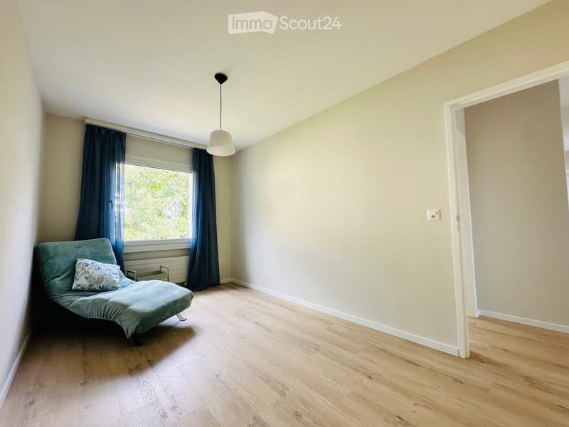 A small, furnished room with light wooden flooring, a single window with dark curtains, a hanging pendant light, and a green sofa with a decorative pillow.