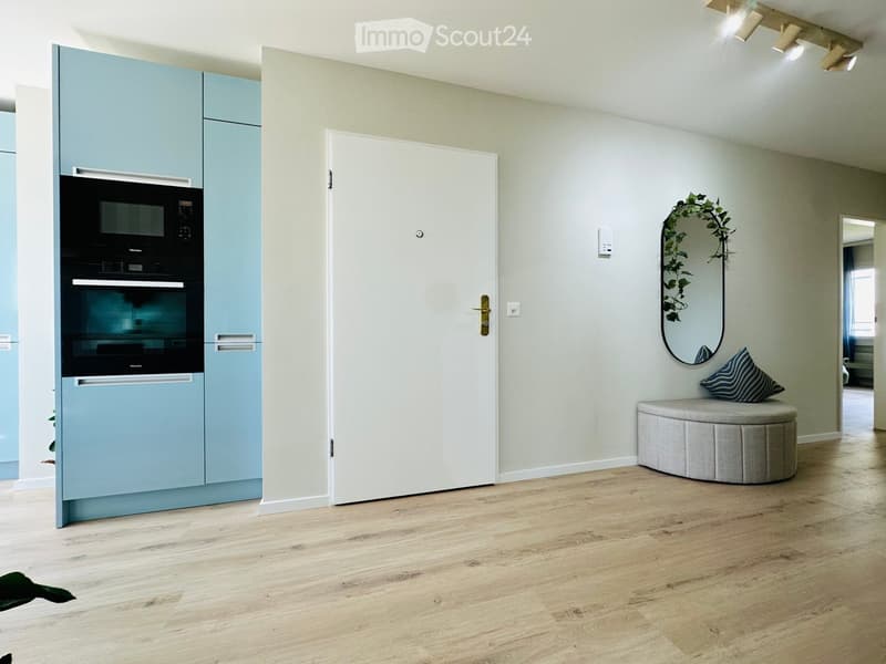 Modern, minimalist room with light blue cabinets, a built-in oven, a round stool with a decorative pillow, and a wall-mounted mirror with a leafy wreath.