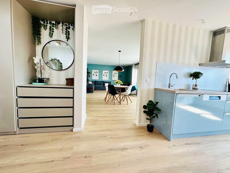 Open-plan living and dining area with a modern kitchen, light blue cabinets, and a wooden dining table surrounded by chairs. The living area features a grey sofa and a round mirror on the wall.