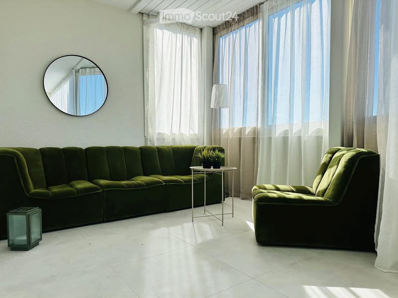 Modern living room with a green sectional sofa, armchair, coffee table, floor lamp, and a round mirror on the wall. The room has large windows with sheer curtains and a small table with plants.