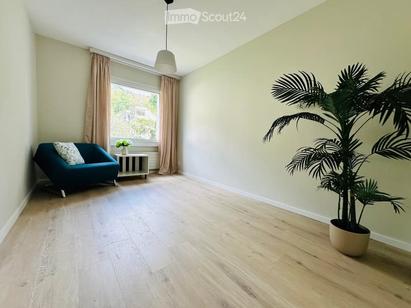 A modern living room with light wooden flooring, a teal sofa, a small white side table with a vase of flowers, and a large potted plant near the window.