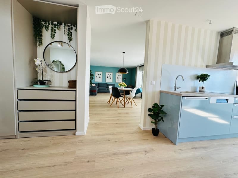 Modern open-plan living and dining area with a kitchen. Features a grey sofa, dining table with chairs, light blue kitchen cabinets, a sink with a faucet, and decorative plants.