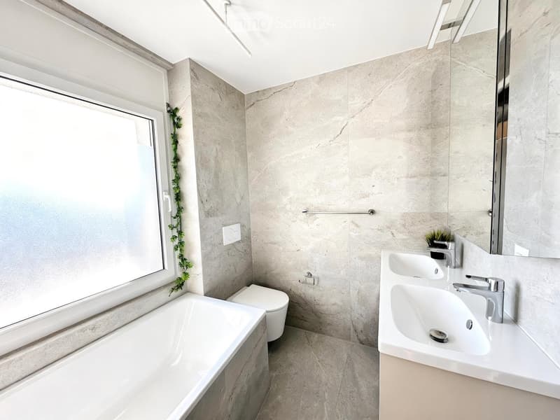 Modern bathroom with a large bathtub, toilet, double sink, and a large mirror. The walls are covered in marble tiles, and there is a window with a decorative plant on the sill.