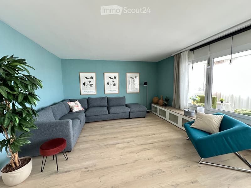 Modern living room with light blue walls, grey sectional sofa, teal armchair, red stool, white side table, and large windows with white curtains.