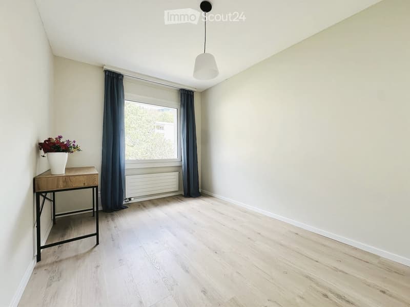 A small, unfurnished room with light wooden flooring, white walls, and a large window with dark curtains. A small wooden table with a vase of flowers sits near the window.