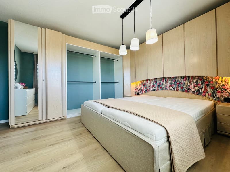 Modern bedroom with a large bed, nightstand, wardrobe, and a floral accent wall. The room features wooden flooring and pendant lighting.