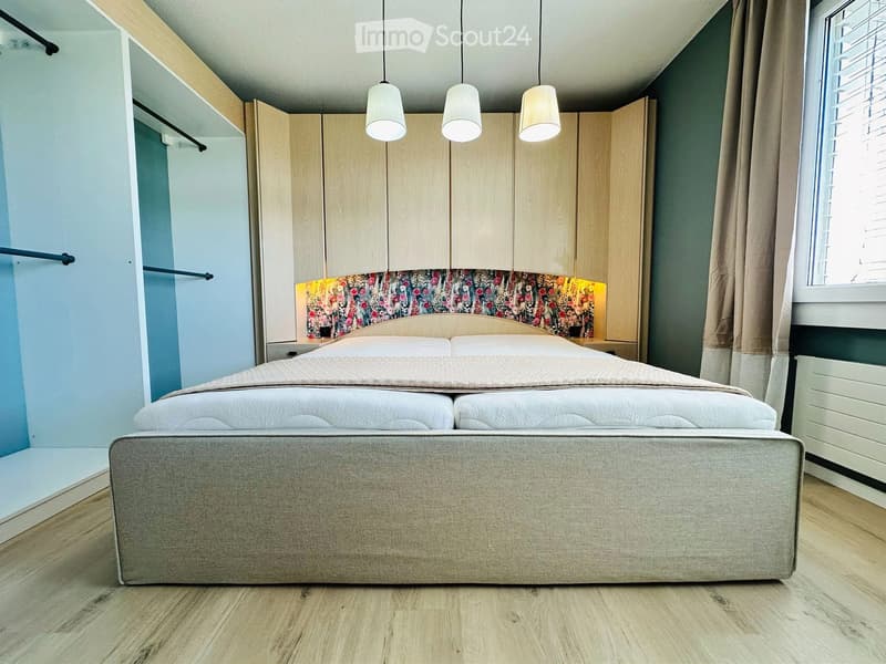 Modern bedroom with a large double bed, built-in wardrobes, and a window with curtains. The room features a floral headboard and pendant lighting.