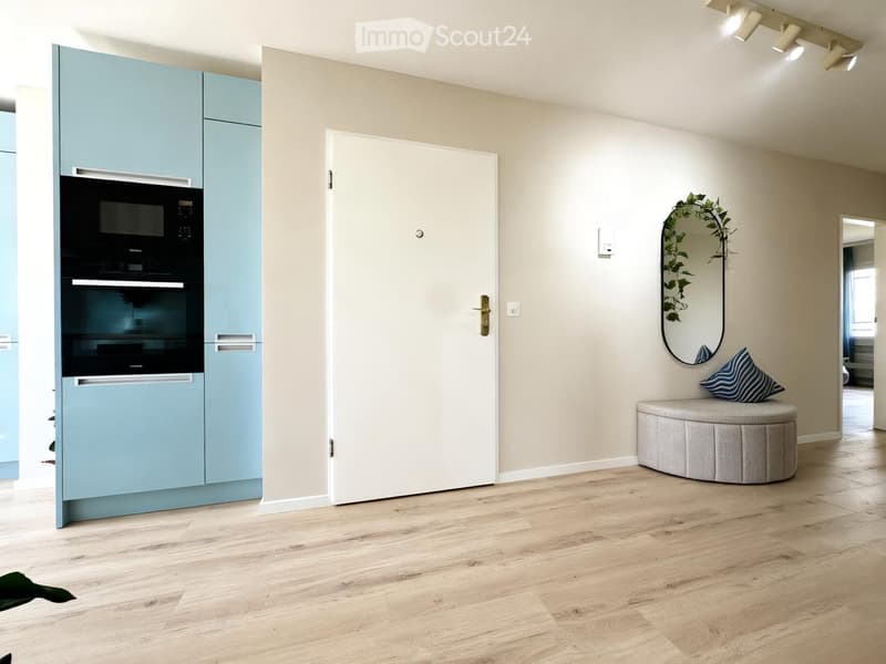 A modern, well-lit room with light blue cabinets, a built-in oven, a round grey stool with a blue and white striped pillow, and a decorative mirror with a leafy plant on it.