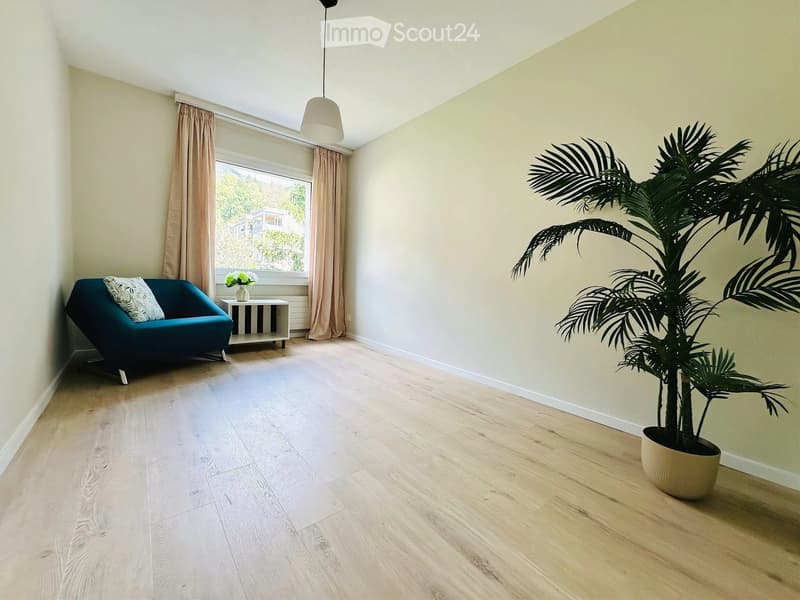 Bright living room with light wooden flooring, a teal sofa, a small side table with a vase of flowers, and a large potted plant near the window.