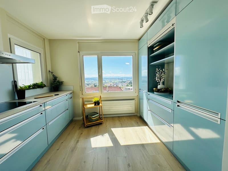 Modern kitchen with light blue cabinets, wooden flooring, and a scenic view through large windows. Includes a countertop with potted plants, a small wooden stool, and a vase on the cabinet.