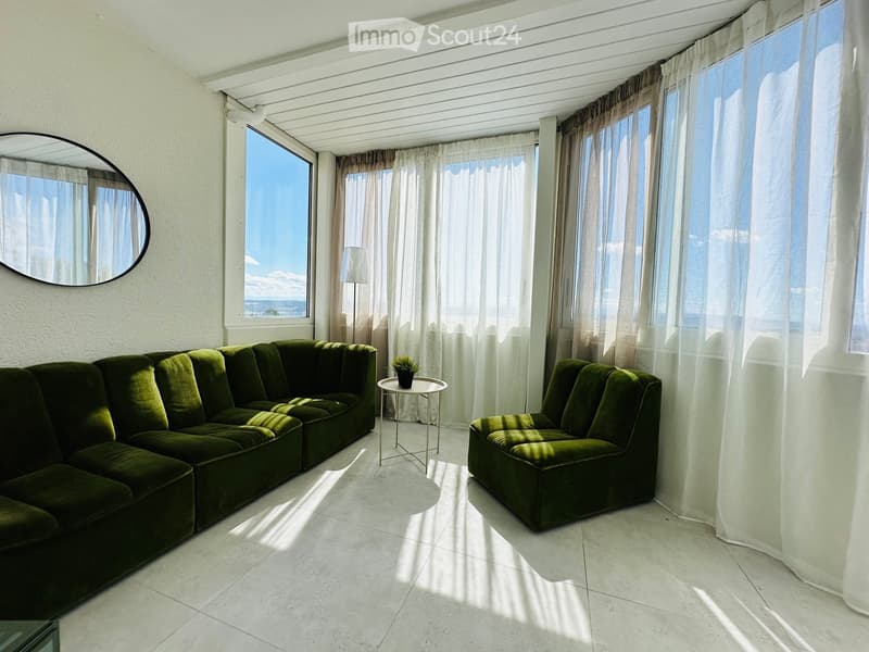 Bright living room with large windows, green sectional sofa, matching armchair, small round table with plant, floor lamp, and a round mirror on the wall.