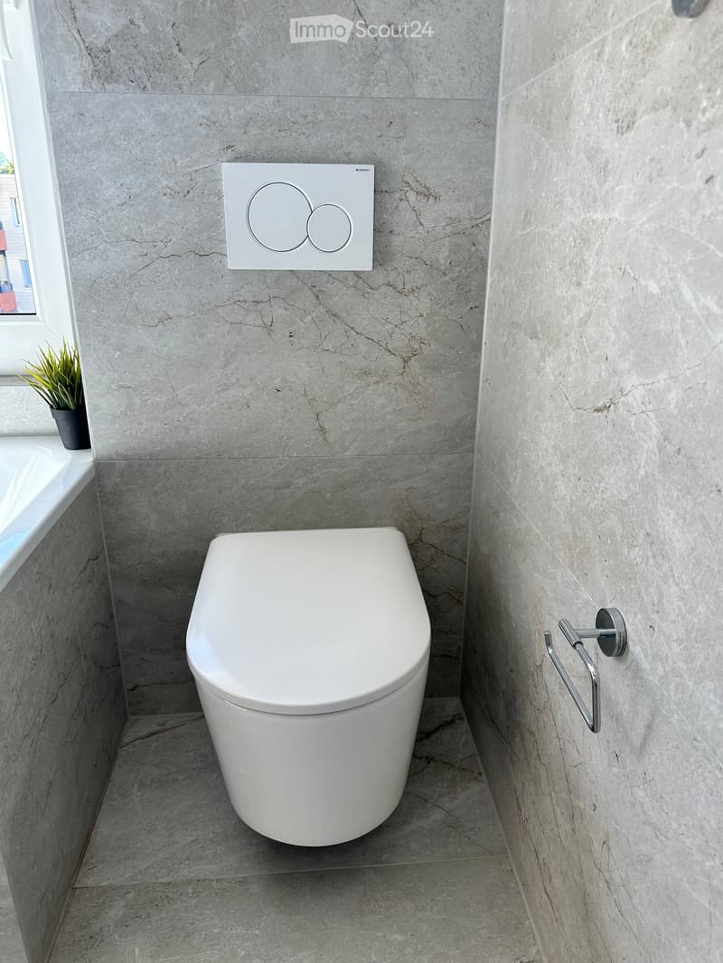 Modern bathroom with a white toilet, marble walls and floor, a bathtub, a control panel, a handrail, and a small potted plant on the windowsill.