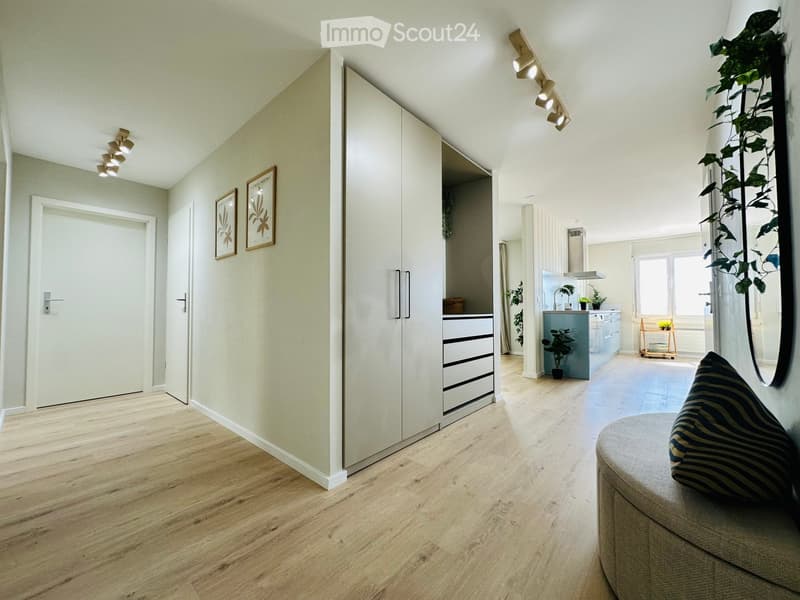 Modern, open-plan living and dining area with light wood flooring, white walls, and contemporary furniture including a sofa, kitchen island, and potted plants.