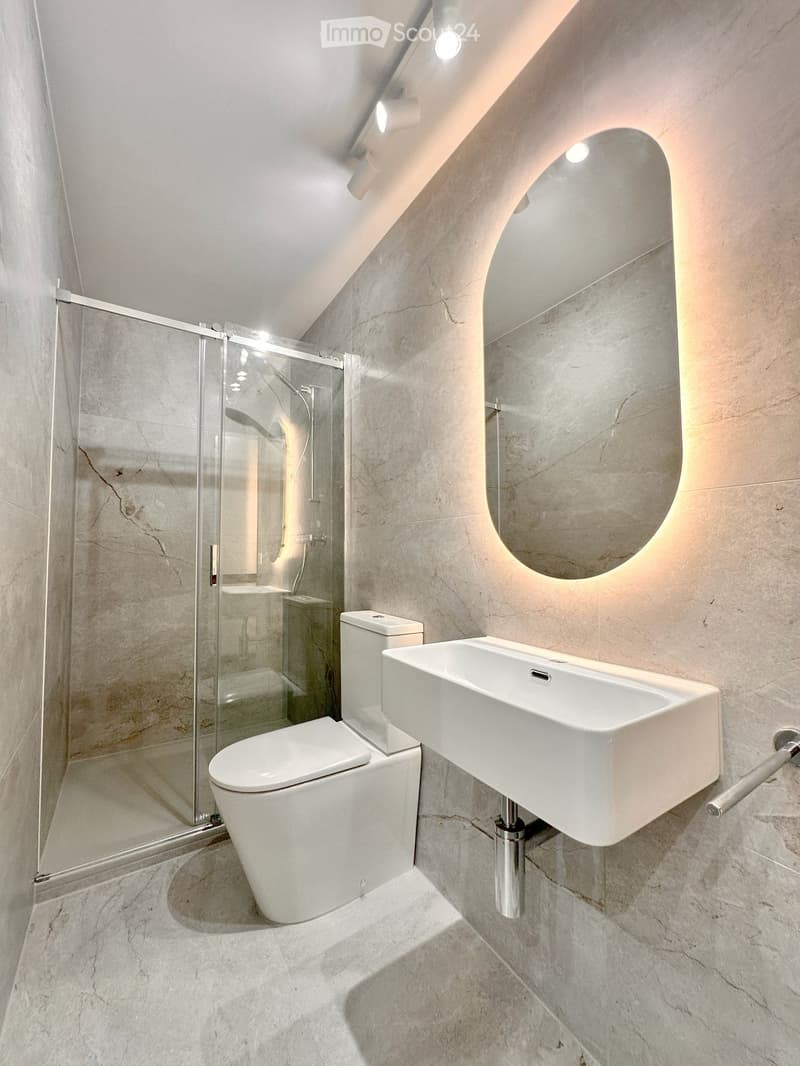 Modern bathroom with marble tiles, white toilet, sink, and a glass-enclosed shower. Features a large, illuminated round mirror and sleek lighting fixtures.