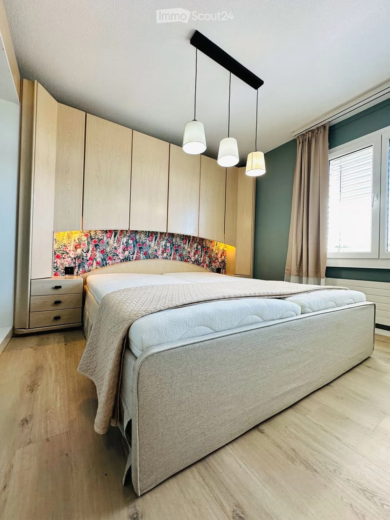 Modern bedroom with a large bed, wooden wardrobe, nightstand, and pendant lights. The room features a floral headboard and a window with curtains.