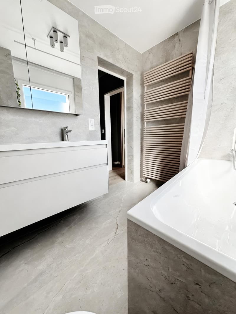 Modern bathroom with a white bathtub, sink, mirror, and wooden radiator. Marble flooring and walls.