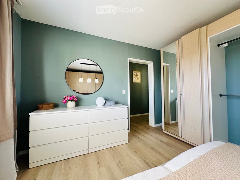 Modern bedroom with light green walls, wooden floor, white dresser, round mirror, and a bed with white bedding.