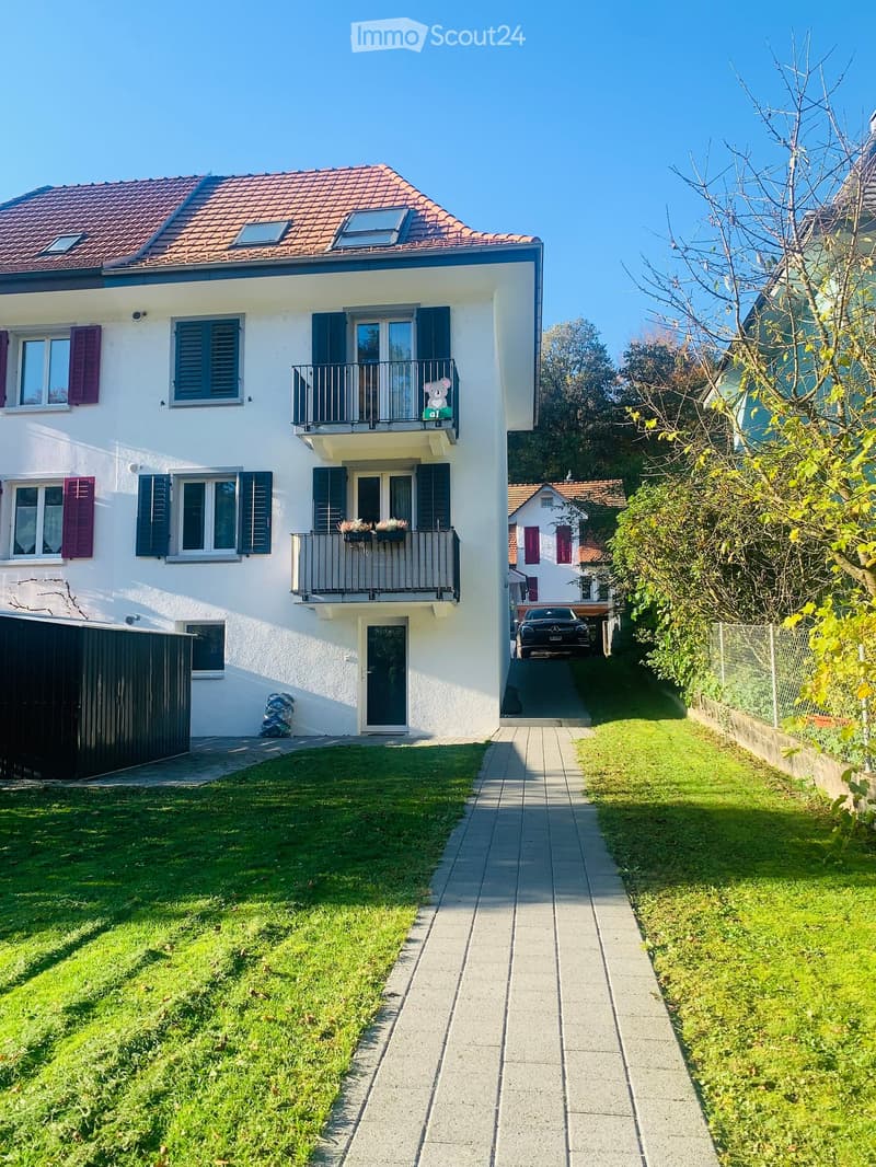 Two-story house with white exterior, red-tiled roof, multiple windows with shutters, balconies, and a small garden area with a pathway leading to the entrance.