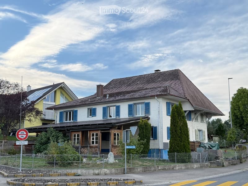 Two-story house with white brick exterior, blue shutters, and a brown tiled roof. Features a covered porch with wooden doors and windows, surrounded by a low fence and greenery.