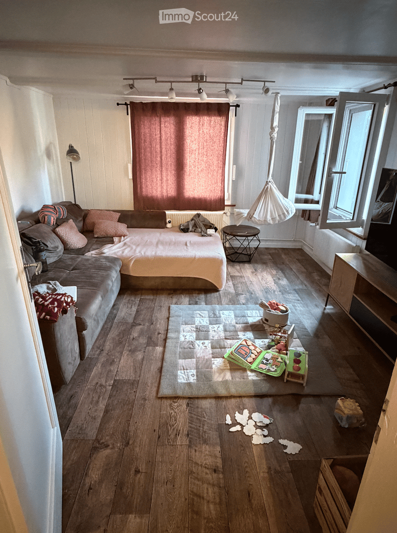 A cozy, multi-functional room with a bed, sofa, small table, and various toys scattered on the floor. The room features wooden flooring, large windows, and a hanging decorative element.