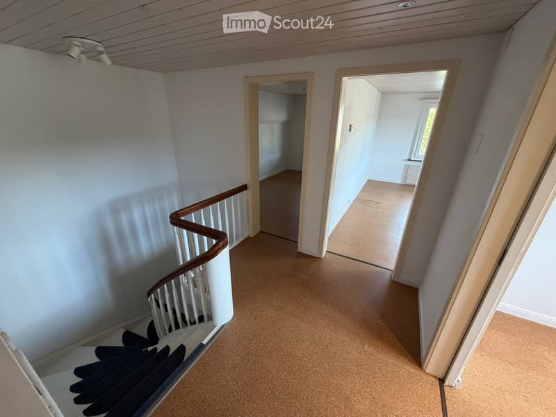Empty hallway with carpeted floor, white walls, and a staircase with wooden handrail leading to another room.