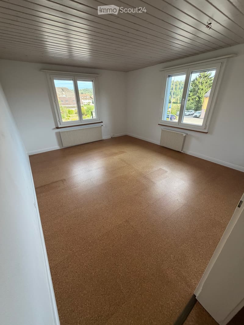 Empty room with wooden flooring, white walls, and two windows offering a view of the outside. Radiators are present under the windows.