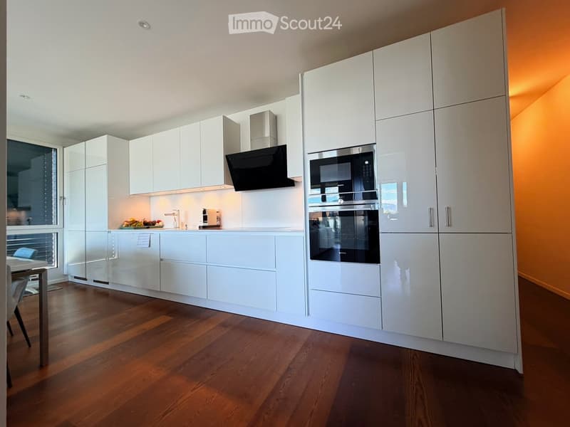 Modern kitchen with white cabinetry, stainless steel appliances, wooden flooring, and a small dining area visible through an open doorway.
