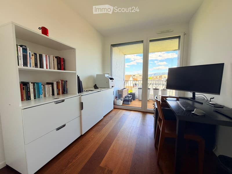 A modern, well-lit study or home office with a large wooden desk, a computer setup, a bookshelf filled with books, and a large window offering a view of the outdoor area.