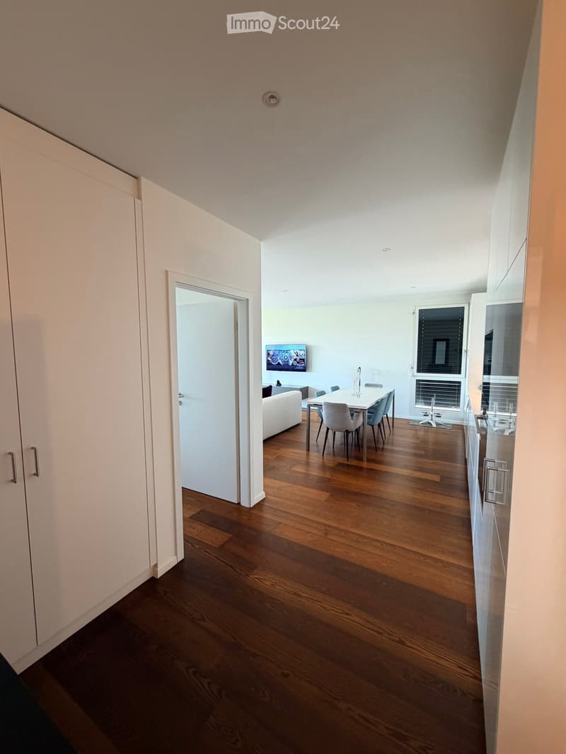 Open-plan living and dining area with wooden flooring, white walls, and modern furniture including a sofa, dining table, chairs, and a television.