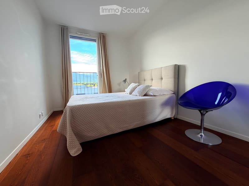 Modern bedroom with wooden flooring, a large bed with a tufted headboard, a purple chair, and a wall-mounted lamp. The room features large windows with curtains, offering a scenic view of the sea and mountains.