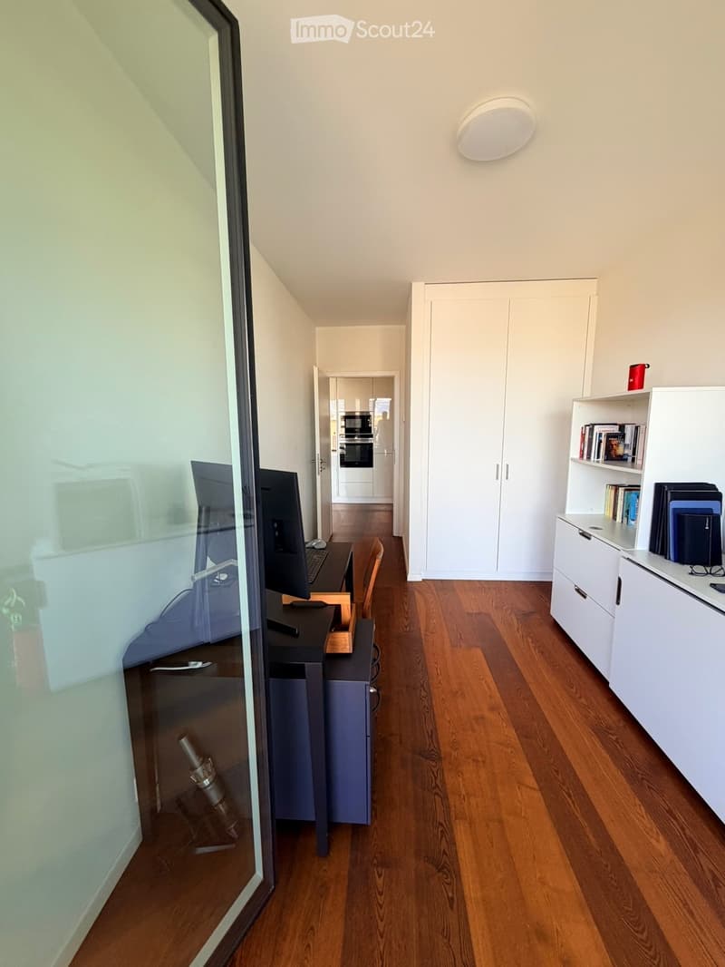 Modern, well-lit room with wooden flooring, a desk setup including a computer, keyboard, and mouse, a bookshelf with books and a cup, and a large white wardrobe. The room features a sleek design with a round ceiling light and a glass partition.