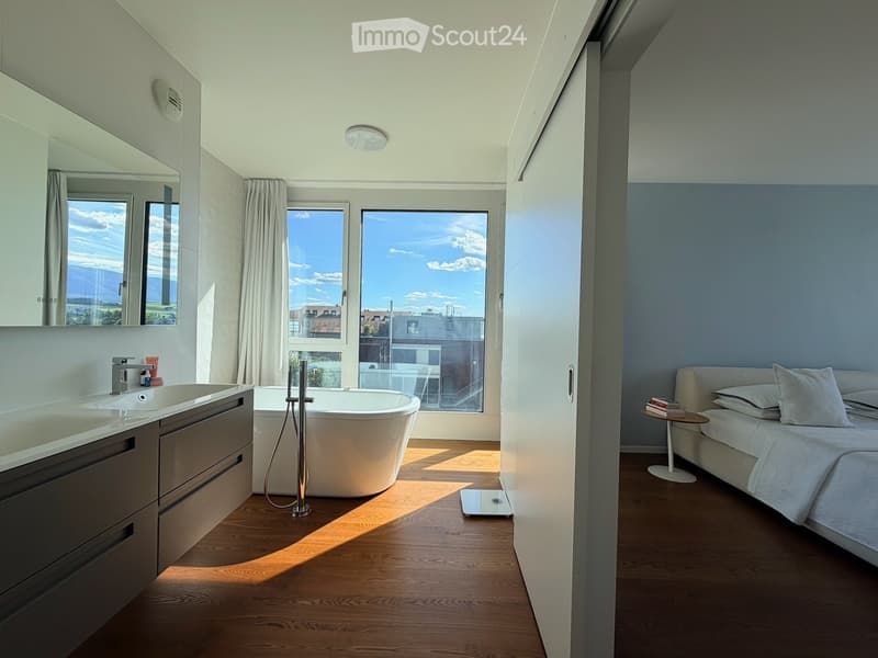 Modern bedroom with a large bed, nightstand, and a separate bathroom area featuring a bathtub, sink, and mirror. Large windows provide natural light and a scenic view.
