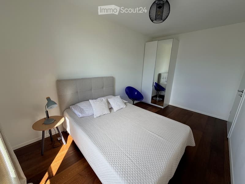 Modern bedroom with a white bed, grey headboard, white pillows, a small nightstand with a lamp, a blue chair, and a large mirrored wardrobe.