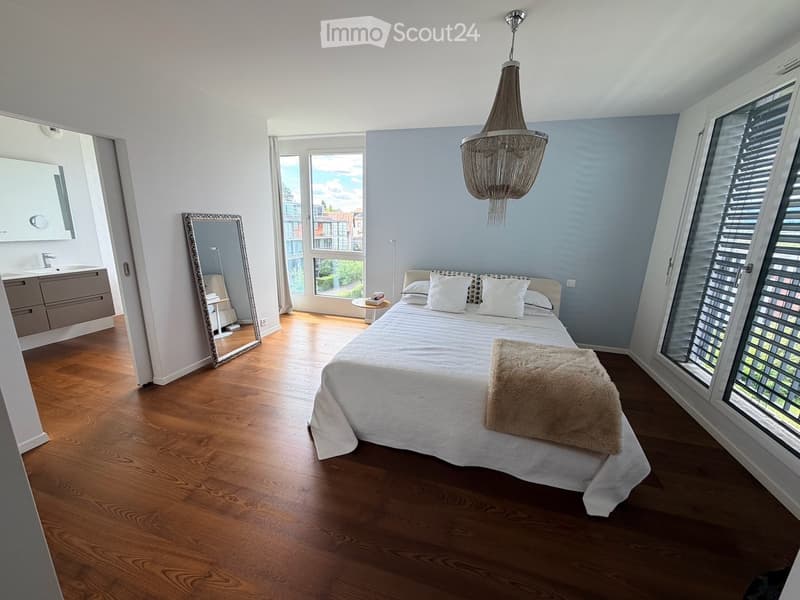 Modern bedroom with wooden flooring, large bed with white linens, a fluffy throw, a large mirror, a small table, and a sink in the adjacent bathroom area.