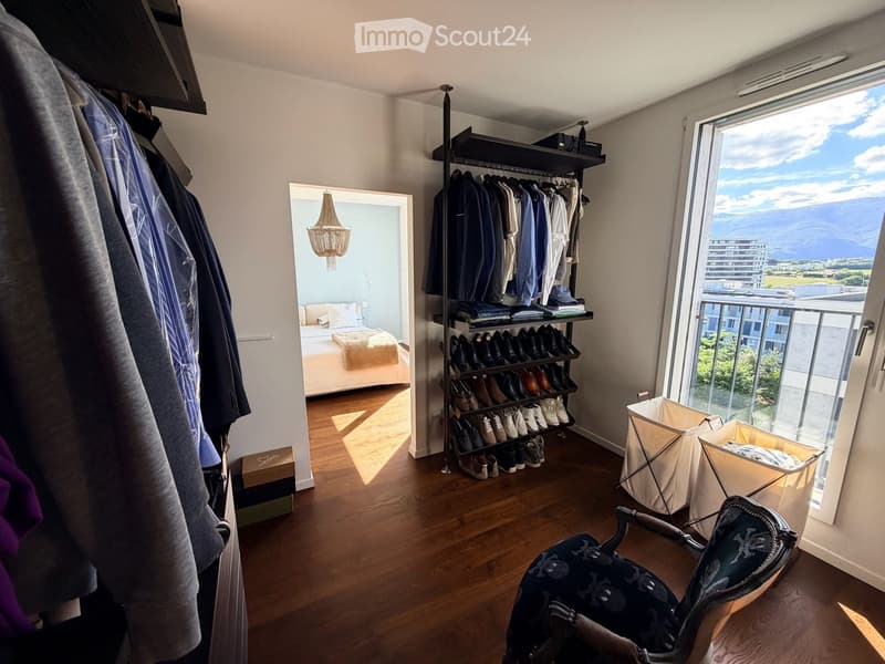 Well-lit walk-in closet with wooden flooring, large window offering a scenic view, and organized storage including a shoe rack, wardrobe, and a small bed visible through an open doorway.