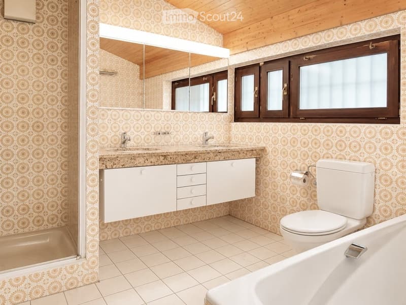Bathroom with a double sink, large mirror, toilet, bathtub, and white cabinets. The walls are covered with patterned tiles, and the floor is tiled. There is a window with wooden frames on the right side.
