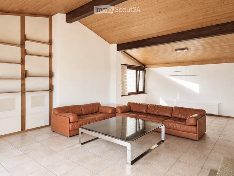 Living room with a brown leather sofa and a glass coffee table, wooden ceiling beams, and a large window with curtains.