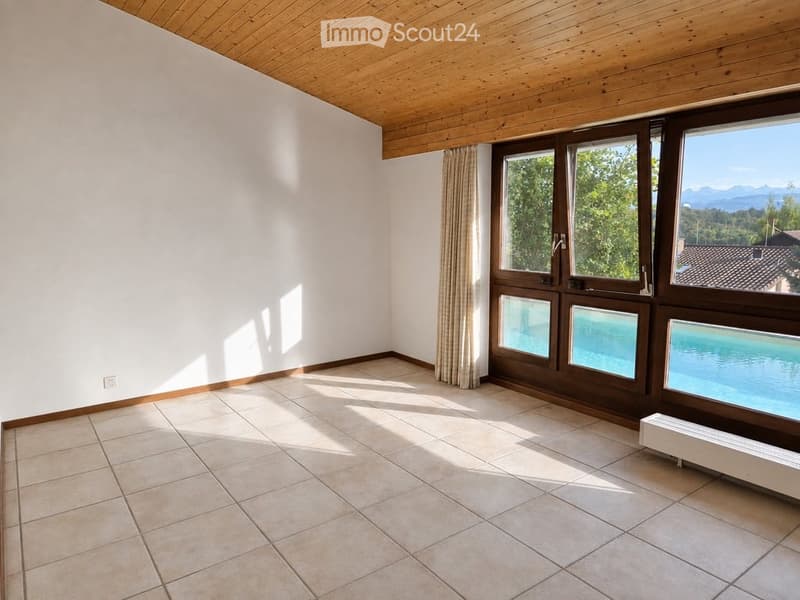 Empty room with tiled floor, white walls, wooden ceiling, large windows with curtains, and a view of greenery and mountains.