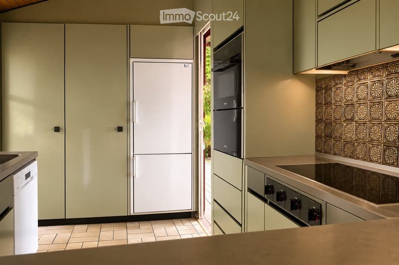 Modern kitchen with light green cabinets, white refrigerator, built-in oven, and dishwasher. Features tiled backsplash and tiled flooring.