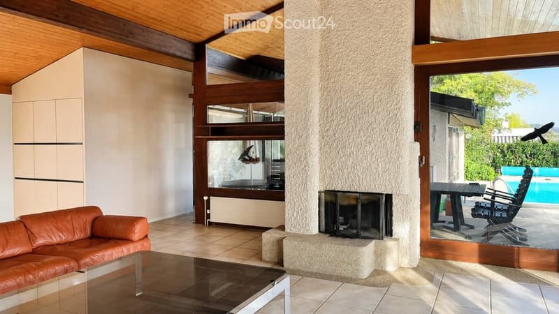 Modern living room with a leather sofa, glass coffee table, and a central fireplace. Large windows offer a view of the outdoor area with a pool and patio furniture.