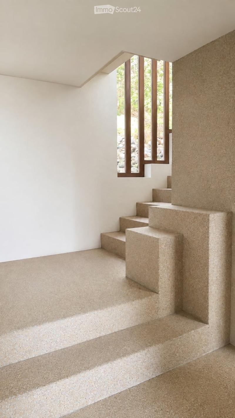 Empty room with white walls, concrete floor, and a small staircase leading up, with a window showing a view of rocks and greenery outside.