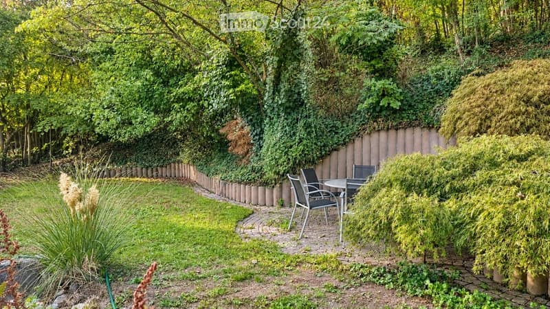 A well-maintained garden with a curved brick pathway, a small seating area with a table and chairs, surrounded by lush greenery and trees.