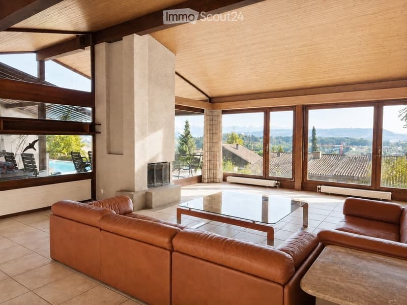 Modern living room with large windows offering a scenic view, featuring a brown leather sofa, a glass coffee table, and a fireplace.