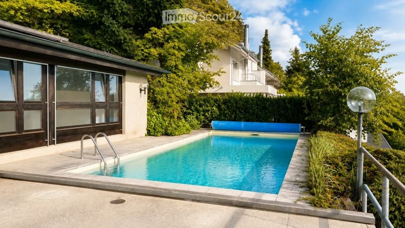Private swimming pool surrounded by greenery, with a modern building in the background and a decorative outdoor lamp.