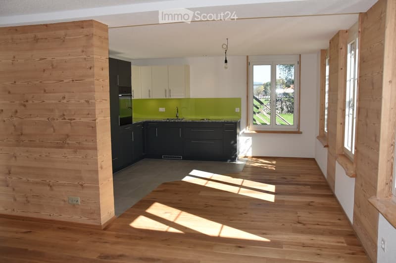 Modern kitchen with light wooden flooring, dark lower cabinets, light upper cabinets, and a green backsplash. Features a window with a view of the garden.