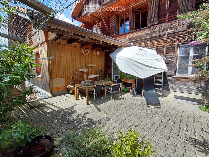 Outdoor patio area with a wooden table, chairs, umbrella, potted plants, and a cat tree near a wooden house.