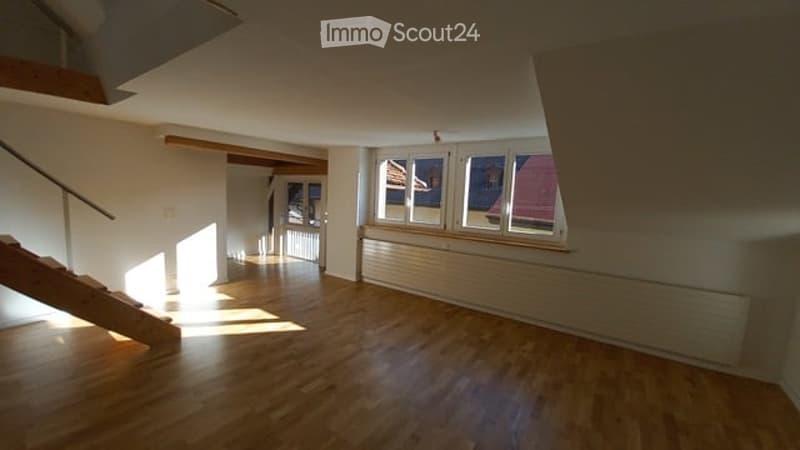 Empty room with wooden flooring, white walls, large windows, and a staircase leading upwards.