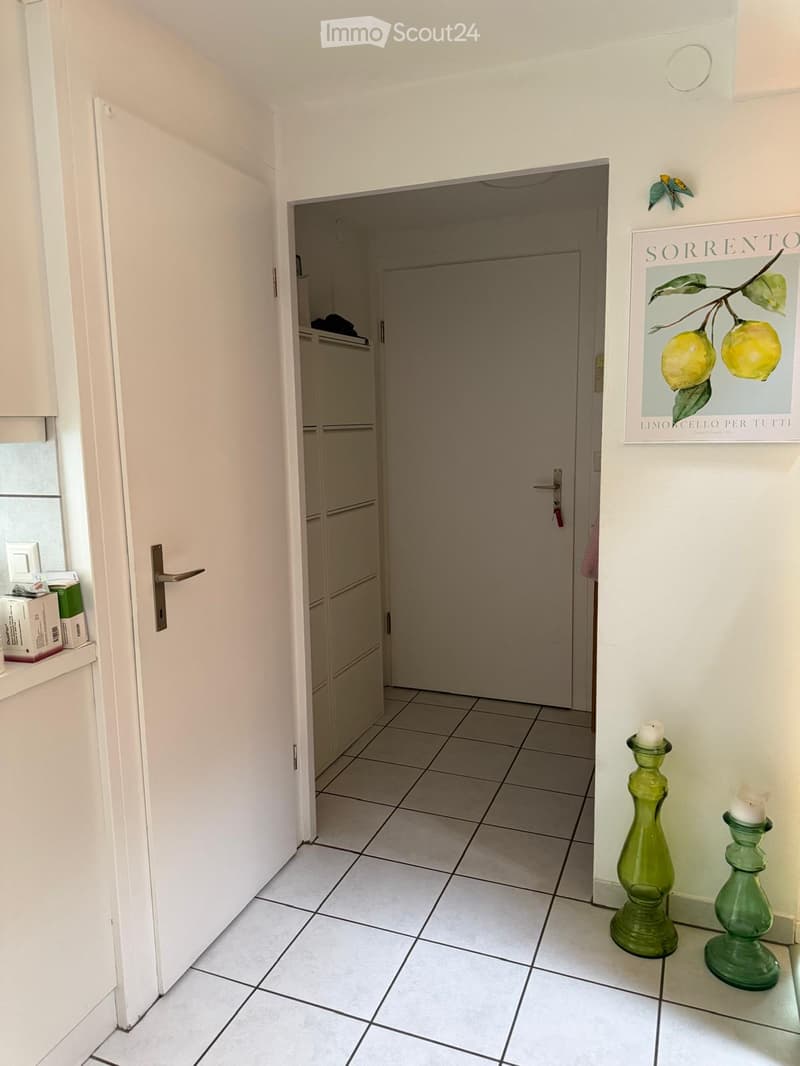 A clean, tiled hallway with white walls, a closed door, and a framed picture of lemons on the wall. Green candle holders are placed on the floor near the wall.