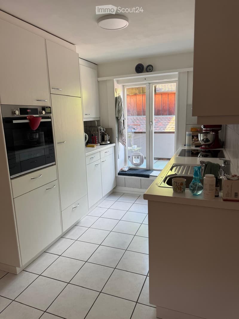 Modern kitchen with white tiled flooring, white cabinets, and stainless steel appliances including an oven and sink. A sliding glass door leads to an outdoor area.