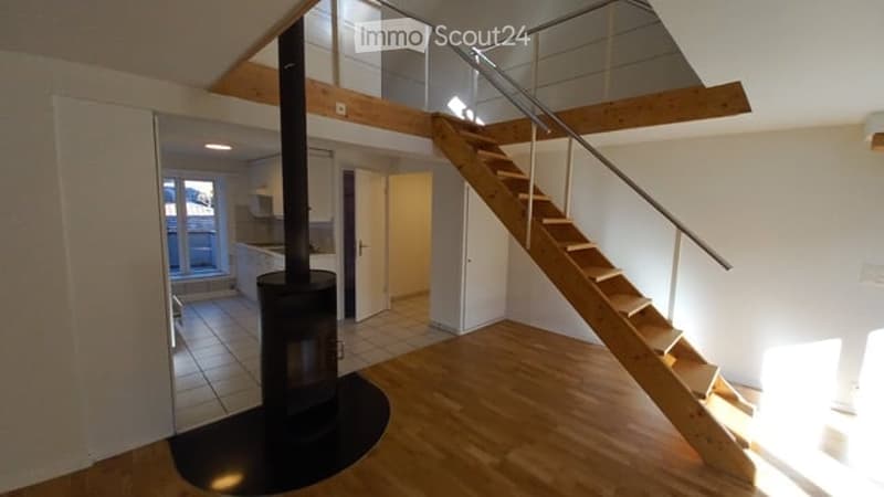 Open-plan living space with a wooden staircase leading to an upper level, a modern black fireplace, and a visible kitchen area with white cabinets and a tiled floor.